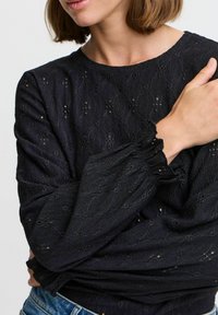 Blouse noire à manches longues en tissu texturé, avec des motifs brodés et des poignets froncés. Encolure ronde et coupe décontractée.