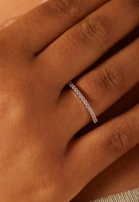 Dünner silberner Ring mit geschwungenem Design, verziert mit kleinen klaren Steinen entlang der Oberseite, vor einem hellbraunen Hautton.