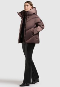 Braune Steppjacke mit hohem Kragen, gesteppter Optik, Reißverschluss vorne und Seitentaschen, kombiniert mit enganliegenden schwarzen Hosen und Stiefeln mit Absatz.