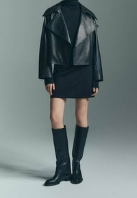 Veste en cuir noir coupée avec des revers larges, col roulé noir ajusté, mini robe et bottes en cuir noir à hauteur de genoux. Textures lisses tout au long.