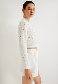 Vit broderad kort blazer med blommönster och långa ärmar, kombinerad med vita högmidjade shorts med rynkat midjeband.