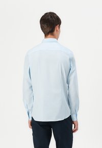 Homme aux cheveux courts et bruns portant une chemise bleu clair à manches longues et un pantalon foncé, debout de dos sur un fond blanc.