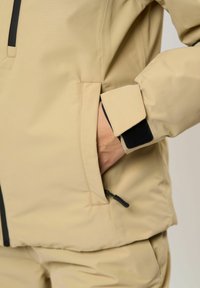 Person in einer beigen Jacke mit einer Hand in einer mit Reißverschluss versehenen Seitentasche, der Jackenärmel hat einen verstellbaren schwarzen Klettverschluss.