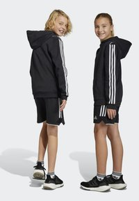 Zwarte hoodie en shorts met witte zijstrepen, gedragen met zwarte sneakers. Kenmerken zijn een kangoeroe-pocket en een sportief ontwerp.