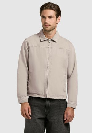 Muž v svetlo béžovej bunde na zips s golierom a čiernych džínsoch stojaci pred jednoduchým svetlým pozadím, mierne pozerá doprava.