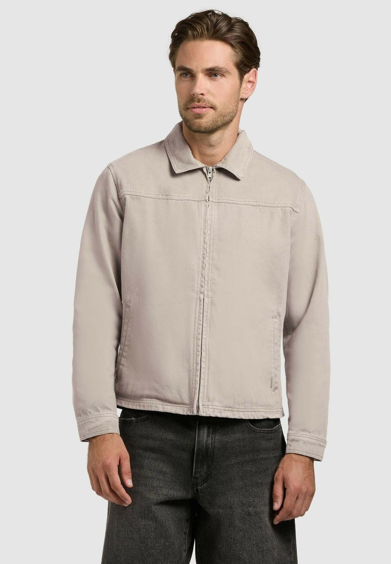 Homme portant une veste zippée beige clair avec col et un jean noir, debout devant un fond clair uni, regardant légèrement vers la droite.