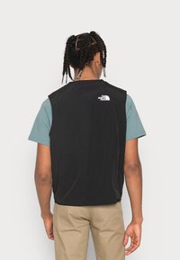 Czarna kamizelka o gładkiej fakturze, z białym logo na górnej części pleców, noszona na niebieskiej koszuli z krótkim rękawem i beżowych spodniach.