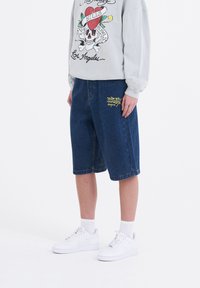 Tummansiniset farkkushortsit, joissa on keltainen "Don Ed Hardy" -logo vasemmassa taskussa. Yhdistetty vaaleanharmaaseen huppariin ja valkoisiin lenkkareihin.