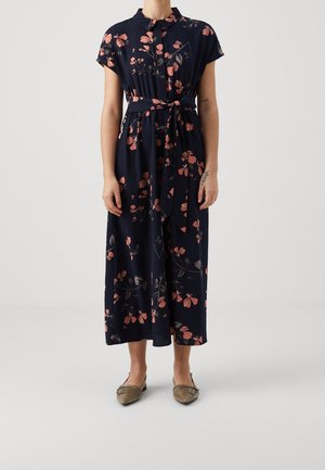 Granatowa sukienka midi w różowy kwiatowy wzór, z krótkimi rękawami, przewiązana w talii, beżowe, spiczaste baleriny na osobie stojącej na białej podłodze.