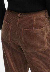 Pantalon en velours côtelé marron avec une texture à côtes verticales, doté de deux poches arrière et d'une taille. Le tissu a une apparence douce et moelleuse.