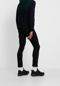 Sweat à capuche bleu marine et vert avec un tissu texturé, jeans noirs ajustés et baskets noires à semelles blanches, stylé avec des manches retroussées.
