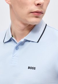 Gros plan d'une personne portant un polo bleu clair avec un col bordé de noir et le logo « BOSS » sur la poitrine.