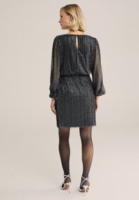 Rochie neagră cu strălucire, cu mâneci lungi din material transparent, talie adunată și detaliu cheie la spate. Asortată cu colanți negri și sandale argintii cu bretele.