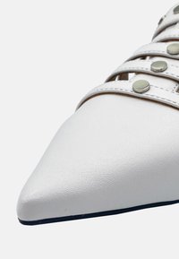 Scarpa sintetica bianca con punta a punta caratterizzata da una superficie testurizzata, design a più cinturini con borchie in tonalità argento e suola nera.