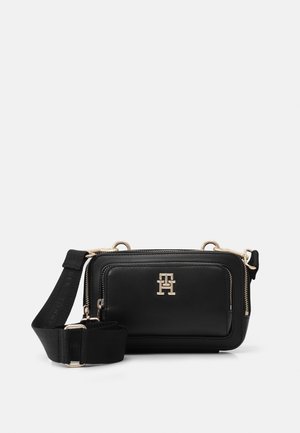 Sac bandoulière noir compact avec poche zippée à l’avant, logo métallique doré et bandoulière en tissu large et réglable sur fond uni.