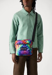 Spalvinga marškinių kriauklė Jansport kuprinė su užtrauktuku, juodais diržais ir logotipu. Suderinta su mėtų žalios spalvos švarku ir rudomis kelnėmis, pabrėžianti drąsų dizainą.