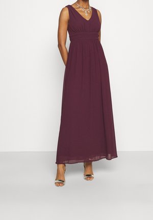 Femme portant une robe longue sans manches d'un violet profond avec un décolleté en V et des talons argentés ouverts, debout devant un fond blanc uni.