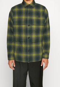 Grön rutig skjorta i texturerad tyg, med två bröstfickor, svarta knappar och krage. Mönstret har gula detaljer.