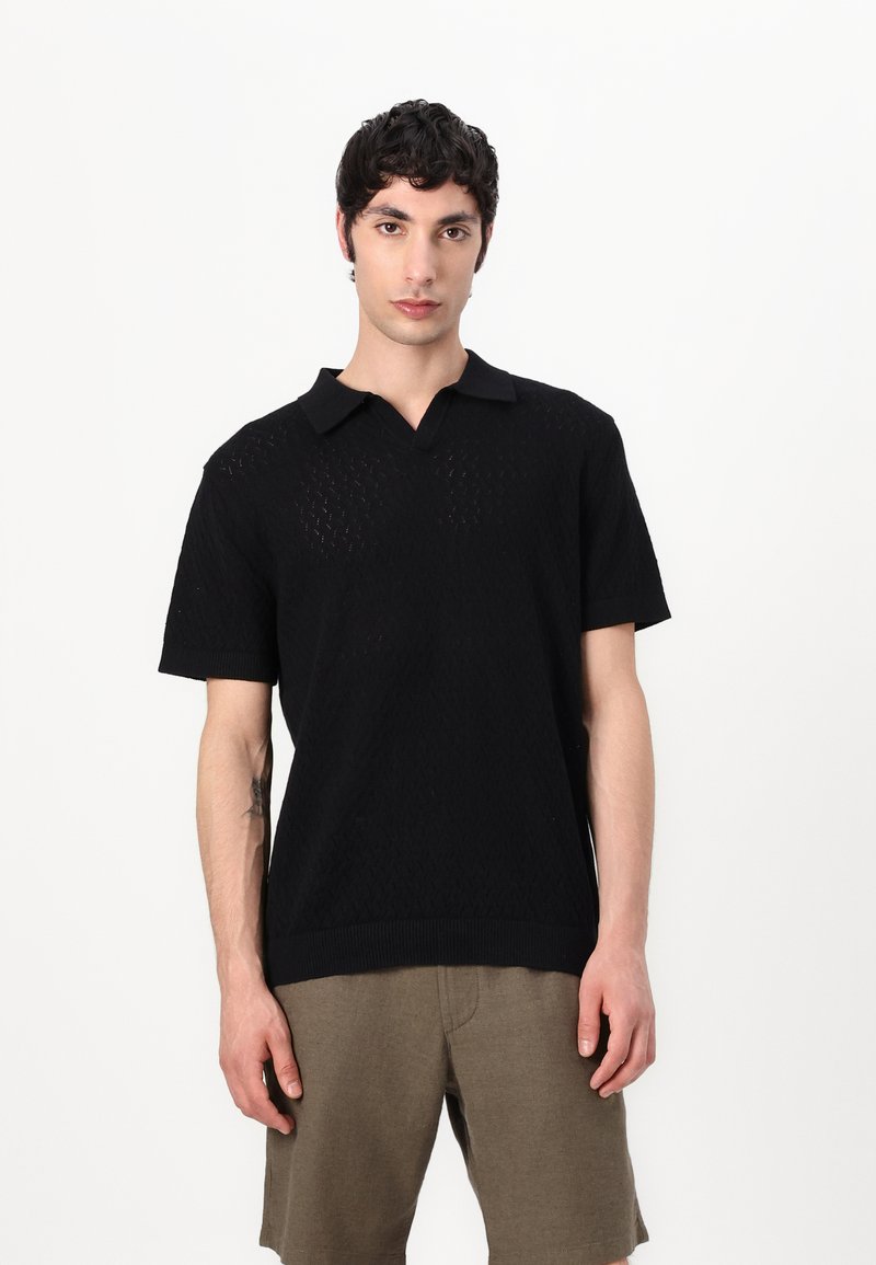 Camisa de polo negra de manga corta, con un patrón texturizado, cuello y puños de canalé, combinada con pantalones cortos caqui.