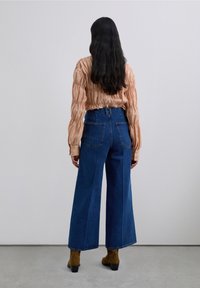Kvinna med långt mörkt hår som vänder sig bort, iförd en persikofärgad rynkad blus, högmidjade vida blå jeans och bruna ankelskots.