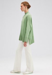 Pulover oversized de culoare verde deschis, cu guler înalt și accente cu model tricotat, purtat cu pantaloni albi evazați și adidași în nuanțe neutre.