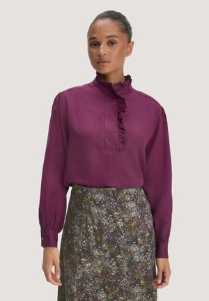 Vrouw die een paarse blouse met lange mouwen en een ruches aan de kraag draagt en een hoge taille bloemenrok, staand tegen een effen achtergrond.