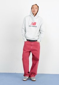 Grauer Kapuzenpullover mit rotem New Balance-Logo, kombiniert mit locker sitzenden roten Hosen und silbernen Sneakers. Einfaches, lässiges Outfit.
