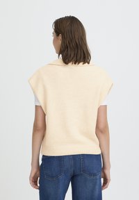 Gilet en tricot pêche clair avec un large col et des manches courtes, porté sur une chemise blanche. Le tissu semble doux, avec un ourlet côtelé.