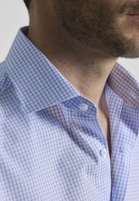 Camisa a cuadros en azul claro y blanco con cuello, que presenta una línea de botones y una tela texturizada. Primer plano del cuello y los detalles de la camisa.