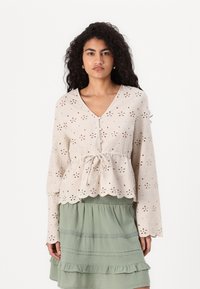 Light beige long-sleeve blouse with eyelet patterns, V-neck, and drawstring waist. Paired with a layered sage green skirt.