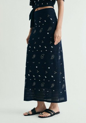 Mujer con falda midi azul oscuro con estampados blancos de paisley y flores, combinada con sandalias planas negras con anillos para dedos.