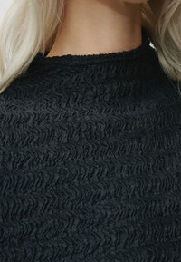 Tessuto nero testurizzato con un motivo ondulato, caratterizzato da un collo alto e una superficie morbida, leggermente stropicciata. Sottolinea la ricchezza del materiale.