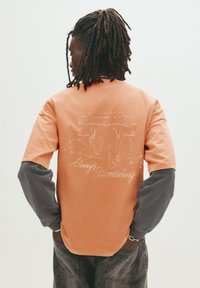 Langærmet skjorte i bomuld med en orange krop og grå ærmer, prydet med et hvidt grafisk design og teksten "Always Wondering" på bagsiden.