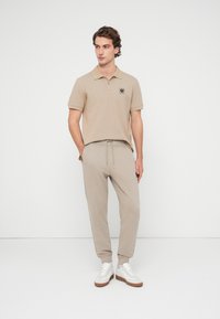 Polo beige à manches courtes avec un écusson logo, assorti à un pantalon de jogging beige avec une taille élastique et des poignets, accompagné de baskets blanches.