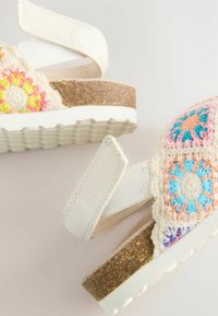 Flerfärgade virkade sandaler med ett vitt justerbart band, en korkfotbädd och en texturerad gummisula. Har blommönster i starka färger.