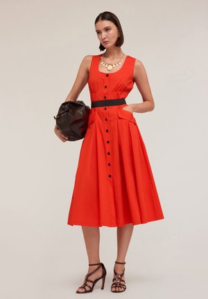Red cotton dress with a fitted bodice, button-down front, pockets, and a black waist belt. Accessories include a brown bag and heeled sandals.