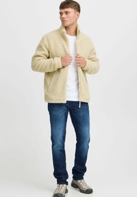Beige fleece jacket with a high collar and front pockets, paired with dark blue jeans and gray shoes, standing against a plain background.