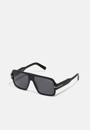 Óculos de sol retangulares pretos com lentes escuras, apresentando um acabamento mate e um detalhe dourado nas hastes. Inclui a marcação nas armas.