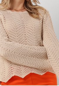 Pull beige en maille avec un tissage lâche et ouvert, ourlet en forme de coquille et larges manches, superposé à un tissu orange vif.