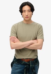Oliven grøn crew-neck t-shirt, korte ærmer, glat tekstur. Blå jeans med et brunt bælte, sort og grønternet flannel bundet om livet.