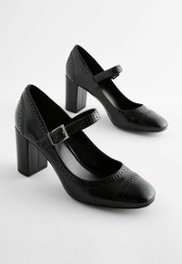 Zapatos de tacón de cuero negro con punta redonda, una correa ancha con hebilla de metal y detalles perforados en la parte superior. Diseño de tacón cuadrado.