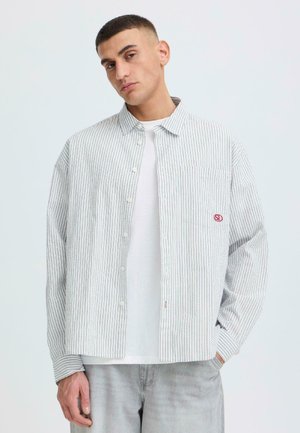 Mężczyzna ubrany w jasno szarą koszulę na guziki z pionowymi paskami, na białym t-shircie i w jasnych dżinsach, stoi z jedną ręką w kieszeni.