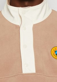 Pullover in pile beige con colletto bianco, dotato di due bottoni a pressione e una toppa rotonda con logo colorato sul petto.