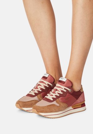 Jambes portant des baskets en daim et mesh marron et rose avec des lacets crème, marquées "Pepe Jeans" sur le côté et la languette, posées sur un fond blanc.