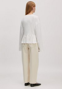 Witte lange mouwen top met een peplum zoom, gecombineerd met crèmekleurige wijde broek en zwarte instappers, gezien vanuit de achterkant.