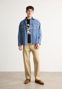 Cămașă din denim de culoare albastră deschisă, tricou negru cu grafică dedesubt, pantaloni bej, pantofi din suede maro. Modelul a pozat în fața unui fundal neutru.