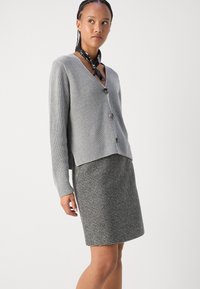 Gilet en tricot gris avec un décolleté en V et trois boutons, associé à une jupe à motifs pied-de-poule noir et blanc. Texture lisse et silhouette ajustée.