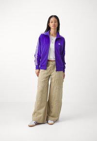 Giacca viola in velluto con zip e strisce bianche, abbinata a pantaloni cargo beige con molteplici tasche e zip, indossata sopra un crop top bianco.