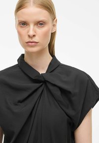 Czarna bluzka z detalem w postaci kokardy przy kołnierzyku. Wykonana z gładkiego materiału, ma szerokie rękawy i luźny krój. Prosty i elegancki design.