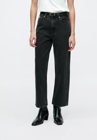 Jeansi negri din denim, talie înaltă, croială dreaptă și textură subtilă, asortați cu o curea neagră și cizme negre cu toc la gleznă.